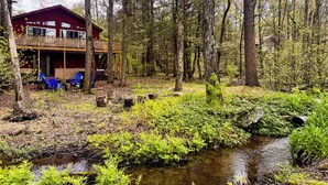 Une cabane dotée d’une terrasse et de meubles de jardin, entourée d’une forêt et bordée par un ruisseau.