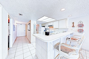 A kitchen with white cabinets, a central island, and a seating area with white chairs. - Coastal Chic Gulf-View 2BR: Suntide III - Unit 604 (South Padre Island)