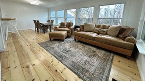 A spacious living room with a large leather sofa, a dining table with chairs, and a window with a view of trees.