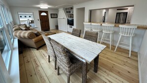 A cozy living room with a wooden dining table and wicker chairs, a sofa with plaid cushions, and a kitchen area with white bar stools and a stainless steel refrigerator.