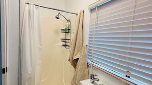 A bathroom with a shower, a sink, and a window with blinds.