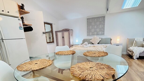 A modern living room with a glass-top dining table surrounded by white chairs, a wooden cabinet, and a large window.