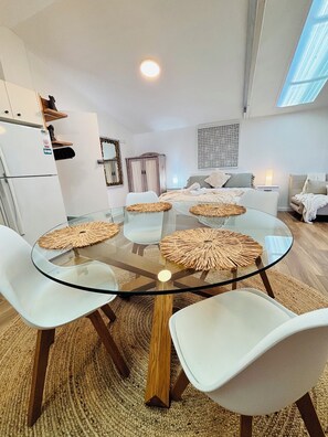 A modern living room with a glass-top dining table surrounded by white chairs, a wooden cabinet, and a large window. - Calm Coastal Hideaway - Cozy, Peaceful, Relaxed & Wellness Inspired (Umina Beach)