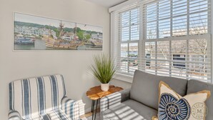 A living room with a large window, a sofa, a striped chair, a small table with a plant, and a framed picture of a harbor scene.