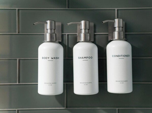 Three white bottles of Brightener Barns body wash, shampoo, and conditioner, each labeled with the product name and size, mounted on a dark tiled wall. - The Mystic River Inn (Groton)