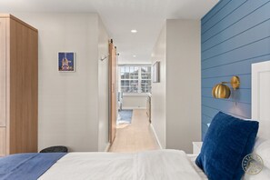 A bedroom with a bed, a blue accent wall, a wooden wardrobe, and a view of a balcony. - The Mystic River Inn (Groton)