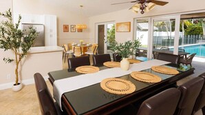 A dining area with a black table, brown chairs, and woven placemats. There is a ceiling fan above, and a view of a swimming pool outside. - Spacious Family Home | Saltwater Pool | Game Room (Palm Beach County)