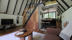Un soggiorno moderno con un tavolo di legno, un divano e una TV. Si vede una scala che conduce a una zona cucina con un bancone e degli sgabelli.
