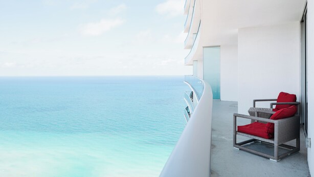 A balcony with a view of the ocean, a wicker chair with red cushions, and a white building. - Oceanfront 36th Floor Condo with Stunning Views Resort and Beach Access (Hollywood)