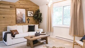 A cozy living room with a white sofa, a wooden coffee table, and a large window with curtains.