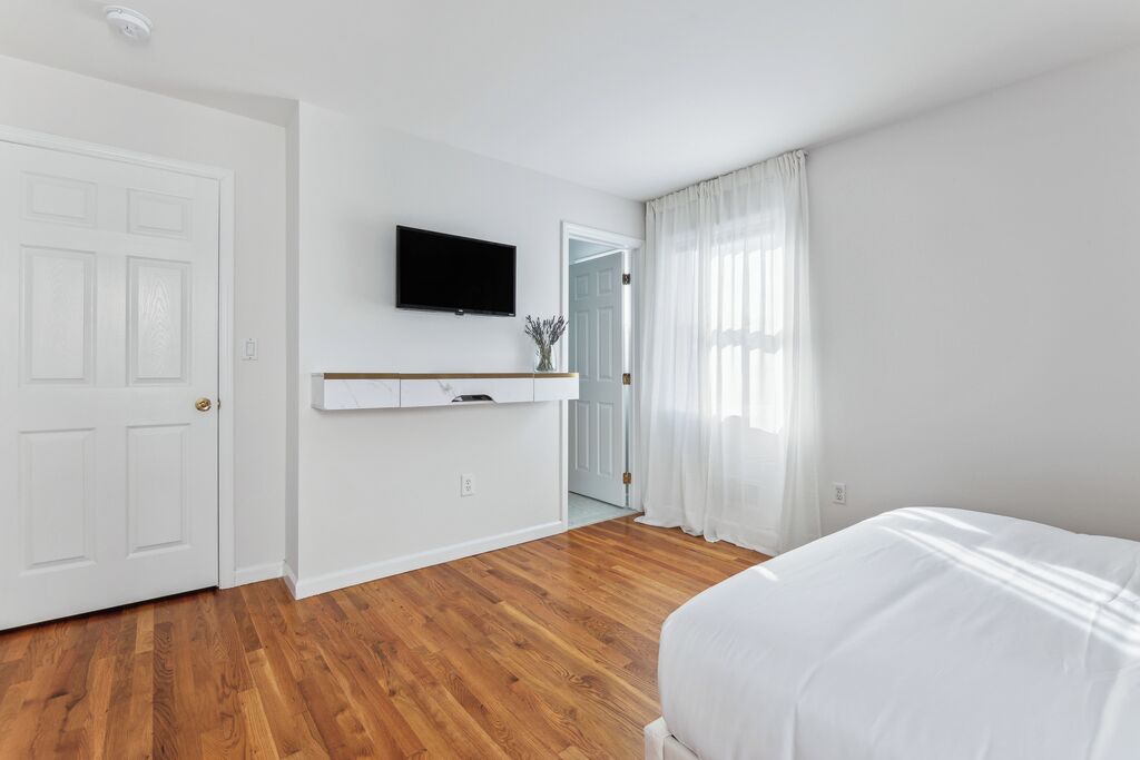A bedroom with a wooden floor, a bed, a TV mounted on the wall, and a door leading to another room.