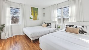 Un dormitorio con dos camas, ropa de cama blanca, un cuadro en la pared y ventanas grandes con cortinas blancas.