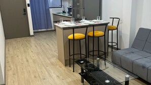 Un espacio habitable compacto con kitchenette, una zona de comedor con taburetes, un sofá y una mesa de centro de vidrio.