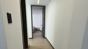 Un pasillo con piso de madera, paredes blancas y una puerta que da a un dormitorio con cama y mesita de noche.