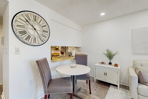 Ein modernes Wohnzimmer mit einem runden Tisch, zwei Stühlen, einer großen Wanduhr, einem weißen Schrank und einer kleinen Pflanze.