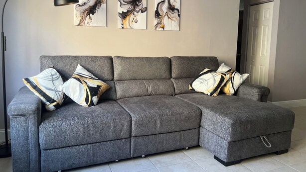 Ein graues Ecksofa mit mehreren Kissen, eine schwarze Stehlampe und drei abstrakte Gemälde an der Wand.