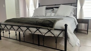 Ein ordentlich bezogenes Bett mit schwarzem Metallgestell, weißer Bettwäsche und einer grünen Tagesdecke. Neben dem Bett steht ein Nachttisch mit einer Lampe.