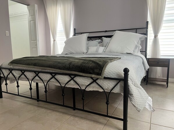 Una cama bien hecha con estructura metálica negra, ropa de cama blanca y un plaid verde. Hay una mesita de noche con una lámpara junto a la cama.