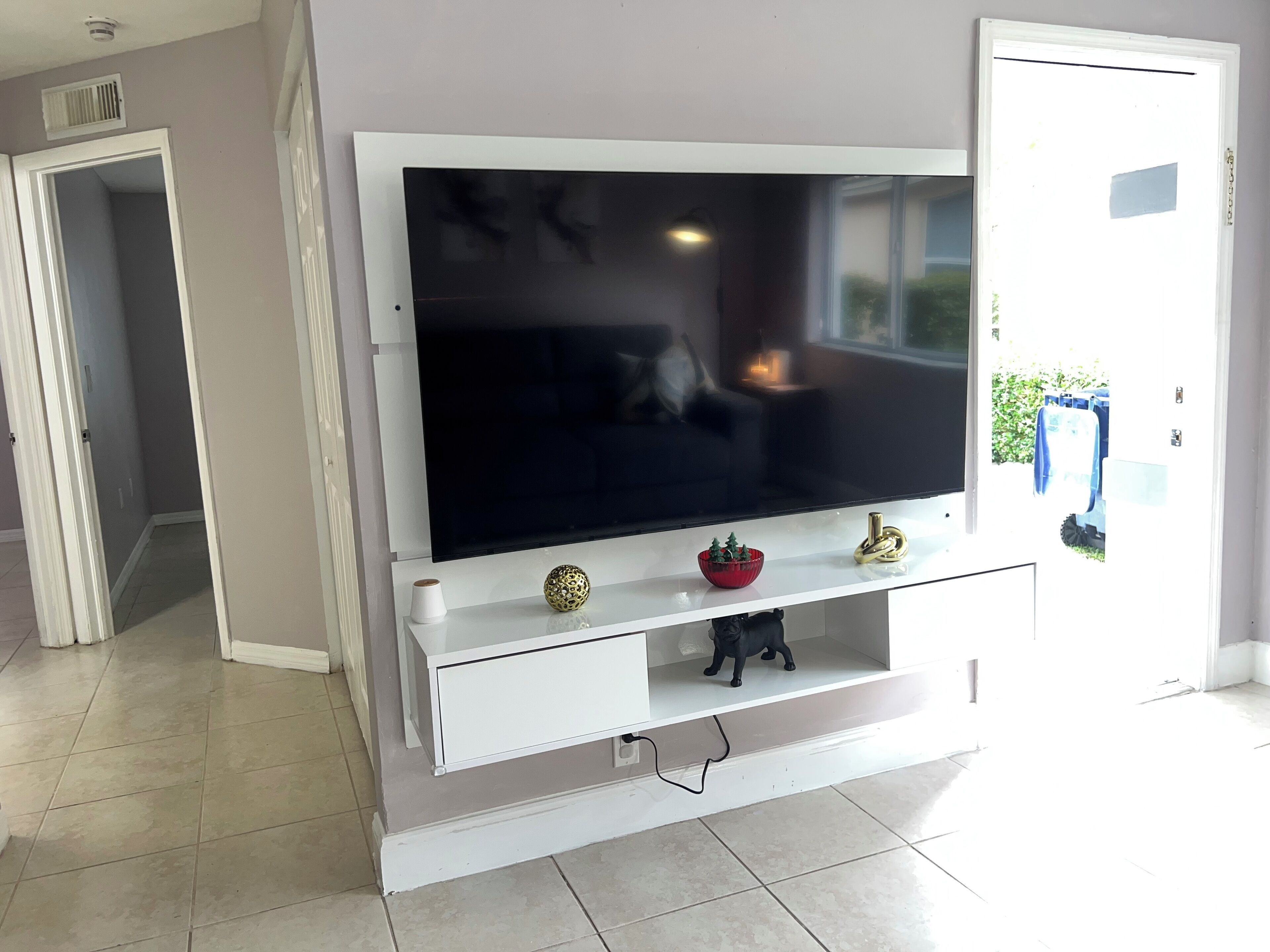 Un salon doté d’un téléviseur à écran plat fixé sur un meuble de rangement blanc, d’une petite étagère ornée d’objets décoratifs et d’une porte donnant sur une autre pièce.