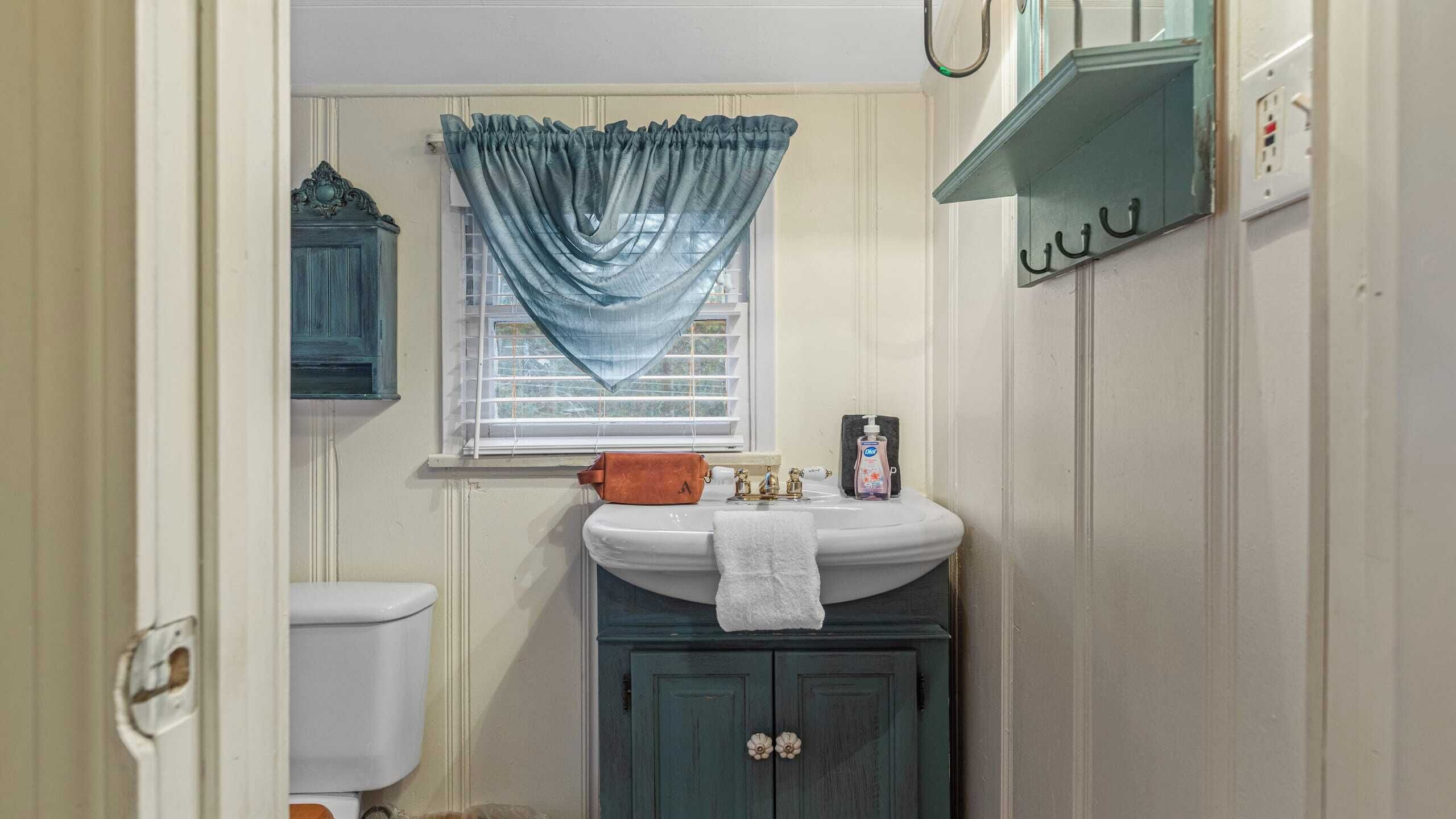 Ein Badezimmer mit Toilette, Waschbecken mit Handtuch, Fenster mit Vorhang und Schrank mit Haken.