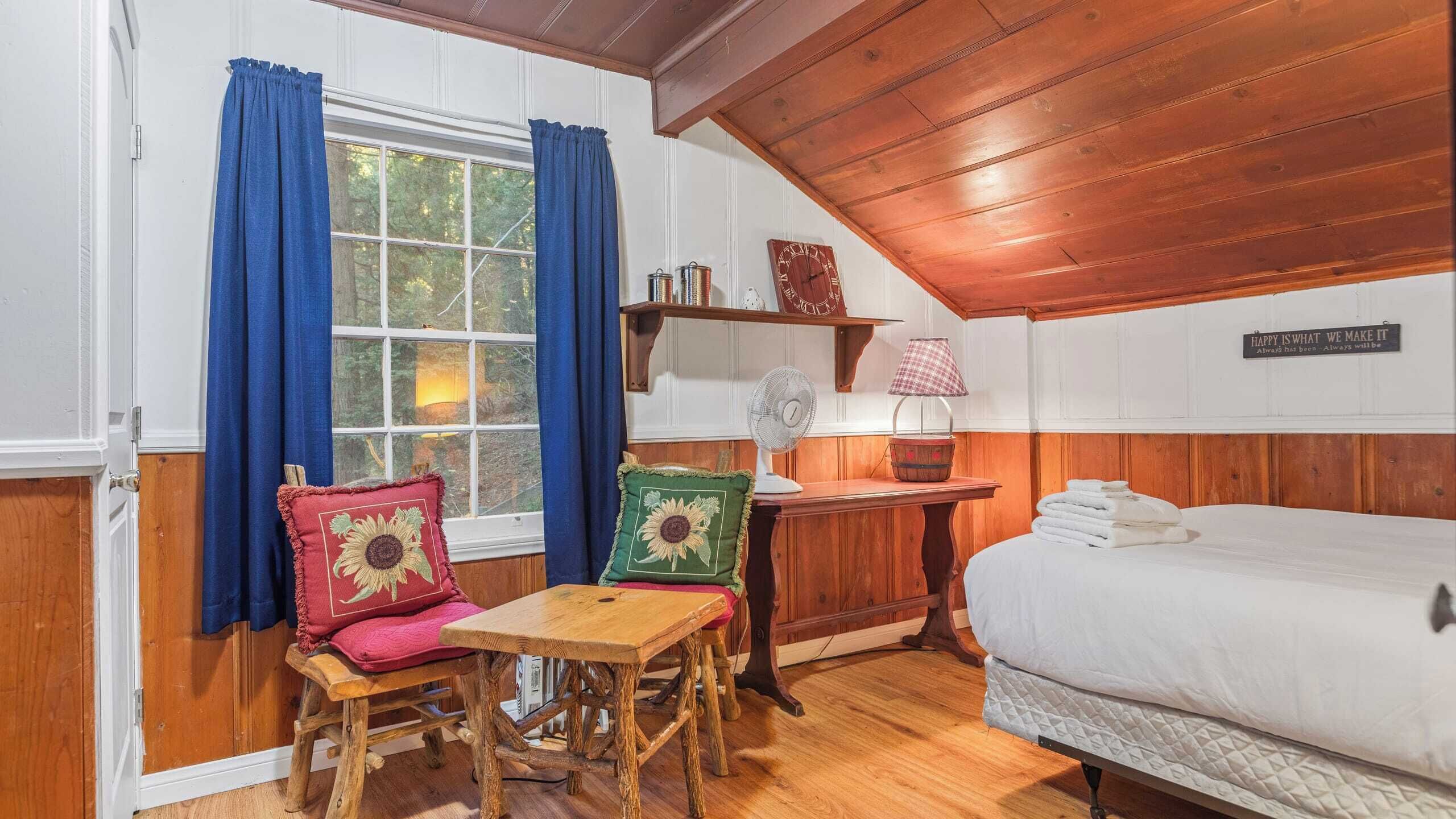 A cozy room with a bed, a wooden table, two chairs, a fan, a lamp, and a shelf with books and a clock.