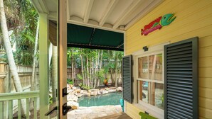 Un patio cubierto con piscina, terraza de madera y vista a un jardín trasero con cerco de madera y plantas tropicales.