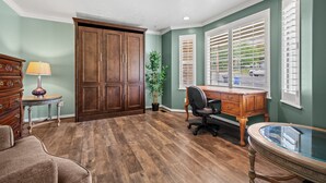 Una habitación con escritorio de madera, una silla, una lámpara, un aparador y un amplio ventanal con contraventanas.