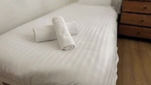 A neatly made bed with a rolled white towel, a wooden nightstand with drawers, and a plain wall.