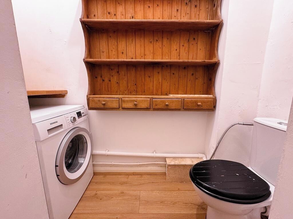 A compact bathroom with a washing machine, a toilet, and wooden shelves above the sink.