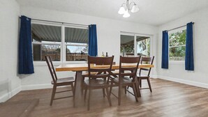 A dining room with a wooden table and chairs, large windows with blue curtains, and a ceiling light fixture. - Spacious 3BR Home Near Lake Hollingsworth (Lakeland)