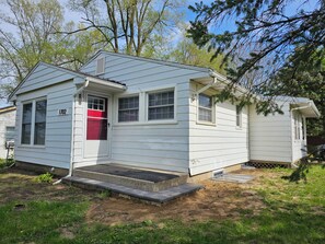 A small, single-story house with a red door and white siding, number 1702, with a small porch and a concrete step leading to the entrance. - Spacious 5-bedroom house in charming Ypsilanti with AC (Ypsilanti)