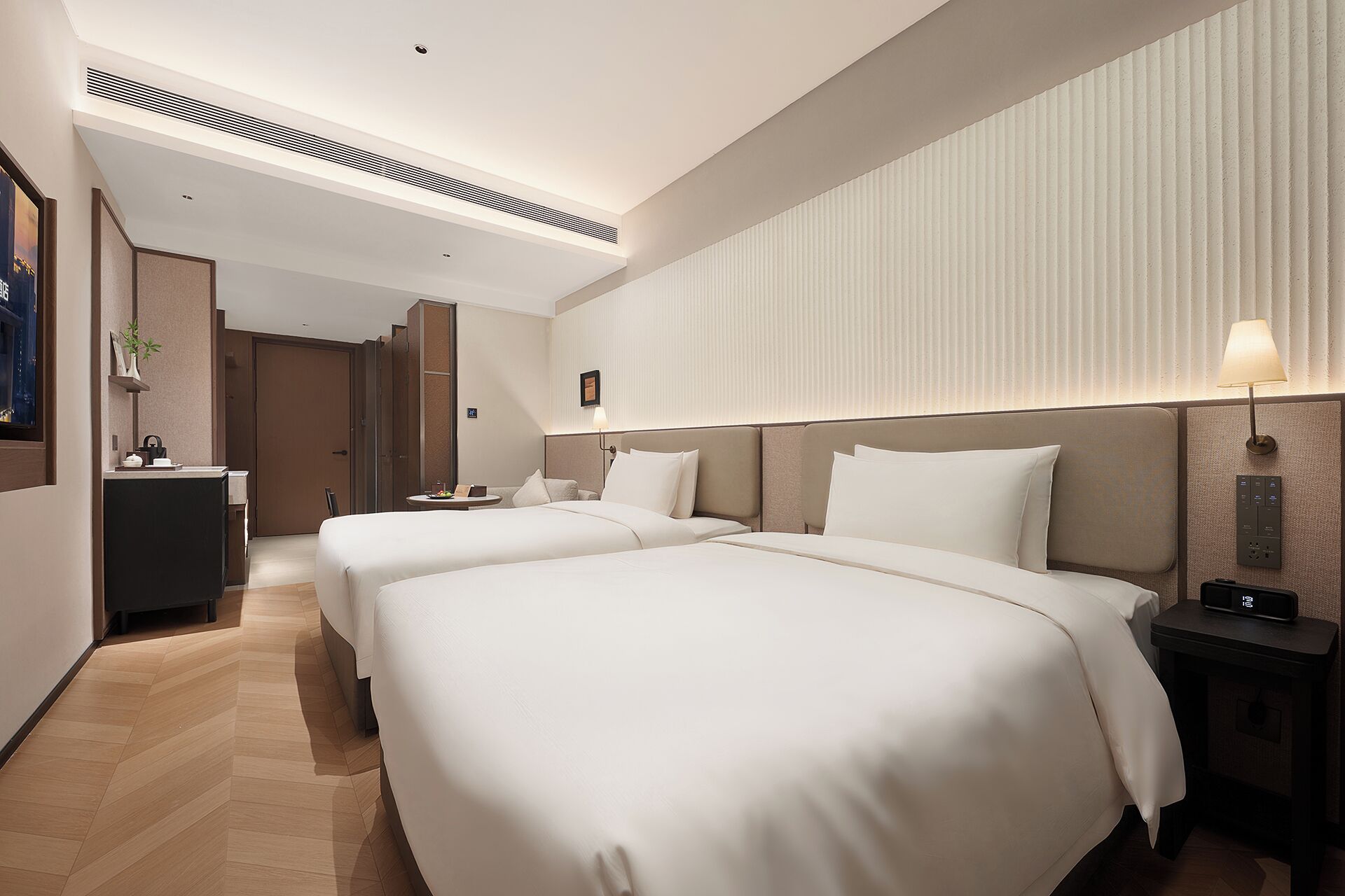 Ein Hotelzimmer mit zwei Betten, einem Schreibtisch, einem Stuhl, einer Lampe und einer an der Wand montierten Bedieneinheit.