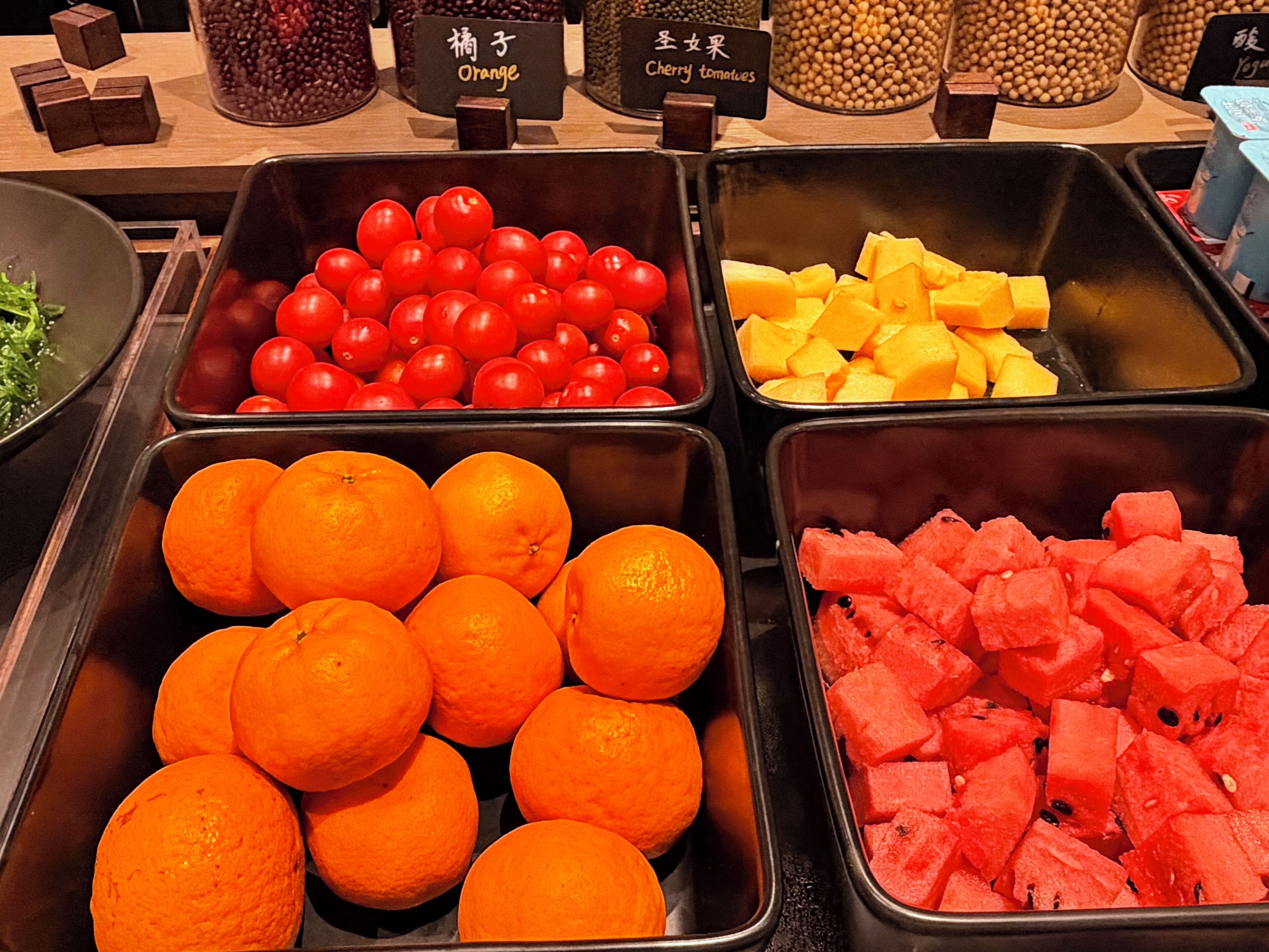 A fruit display with labeled containers of oranges, tomatoes, mangoes, and watermelon.