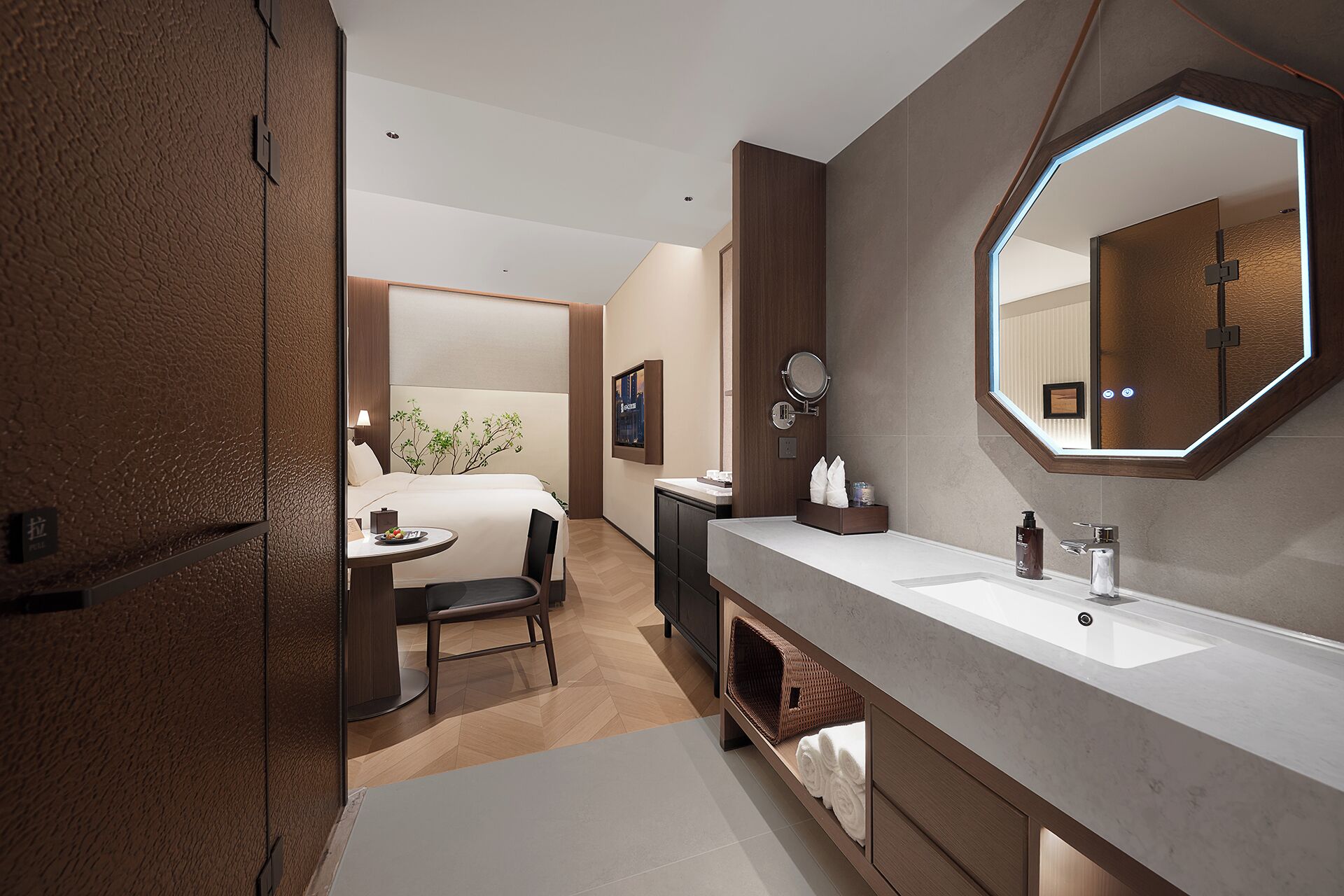 Un moderno baño de hotel con un espejo grande, un lavabo y una tocador. Se ve una pequeña mesa con una silla y una planta al fondo.