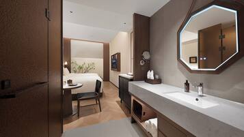Un moderno baño de hotel con un espejo grande, un lavabo y una tocador. Se ve una pequeña mesa con una silla y una planta al fondo.
