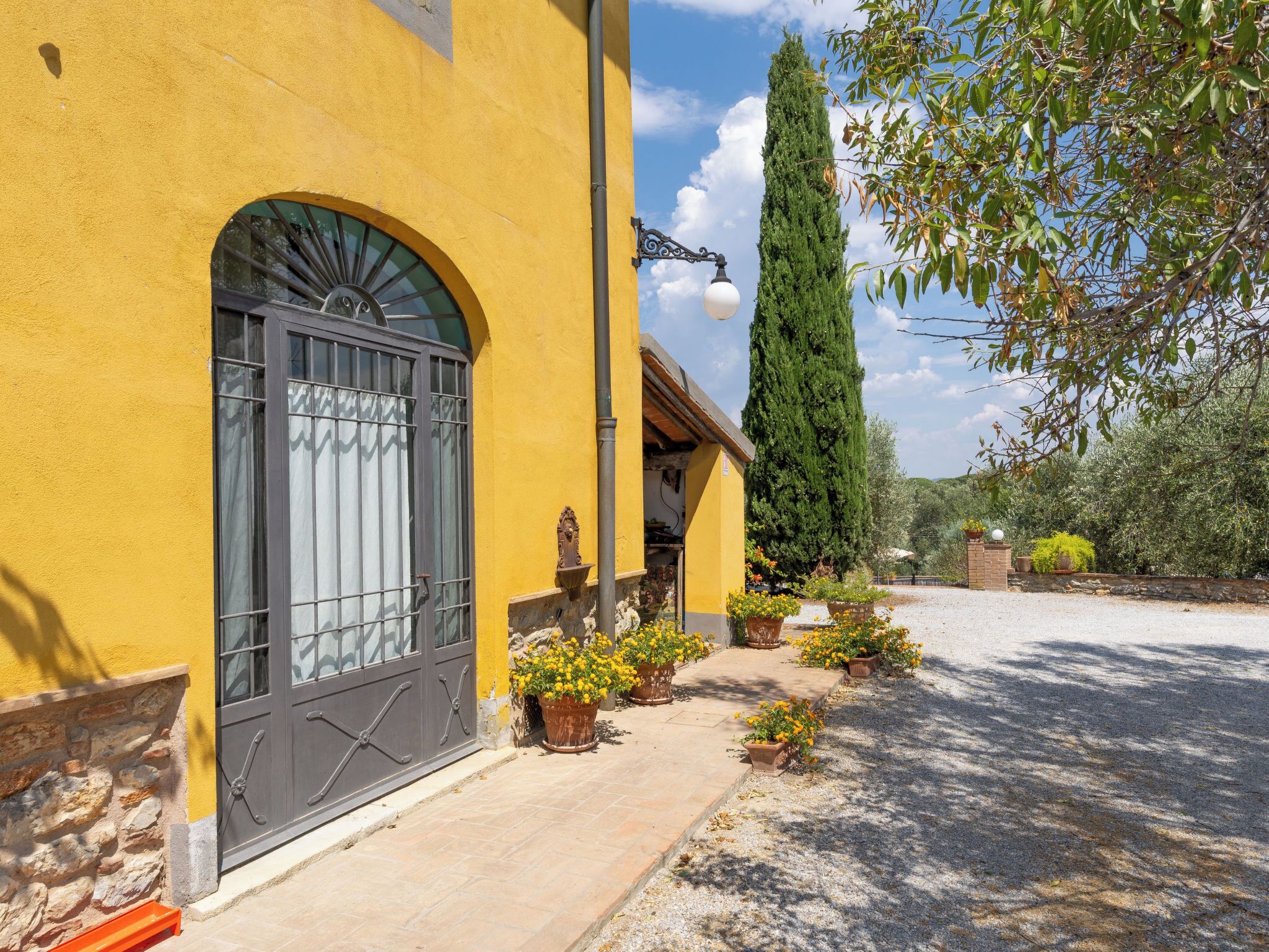 Un edificio amarillo con un amplio ventanal de arco y una pared de piedra. Hay plantas en macetas y un árbol alto en la parte delantera.