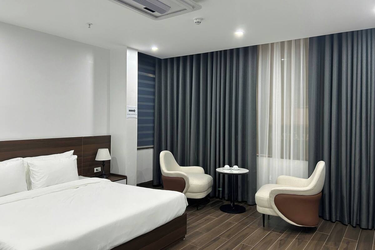 A hotel room with a bed, two armchairs, a small table with cups, and a window with curtains.