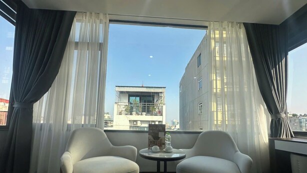 A room with two white armchairs and a small round table, a window with sheer curtains, and a view of a building across the street. - Elegant Hotel (Hanoi)