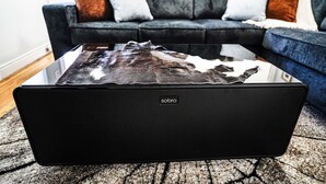 Um alto-falante portátil preto com a marca 'sobro' sobre um tapete texturizado em uma sala de estar.