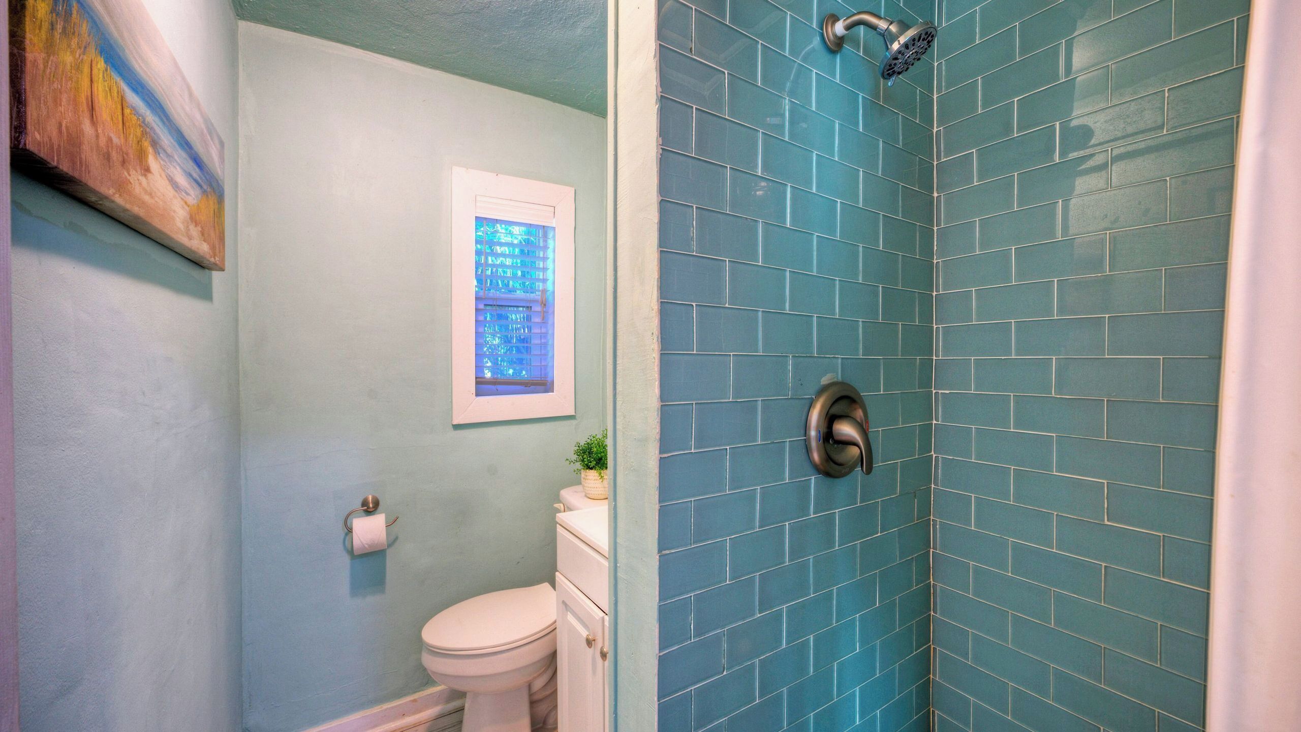 A bathroom with a toilet, a shower with blue tiles, and a framed picture on the wall.