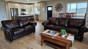 A spacious living room with two leather sofas and a wooden coffee table, a kitchen with white cabinets and modern appliances, and a large window offering a view of the outdoors. - Modern home on acreage with pond, trails, rolling hills, and sunset views (Winterset)