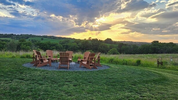 A grassy outdoor area with Adirondack chairs and a table, surrounded by a natural landscape. - Modern home on acreage with pond, trails, rolling hills, and sunset views (Winterset)