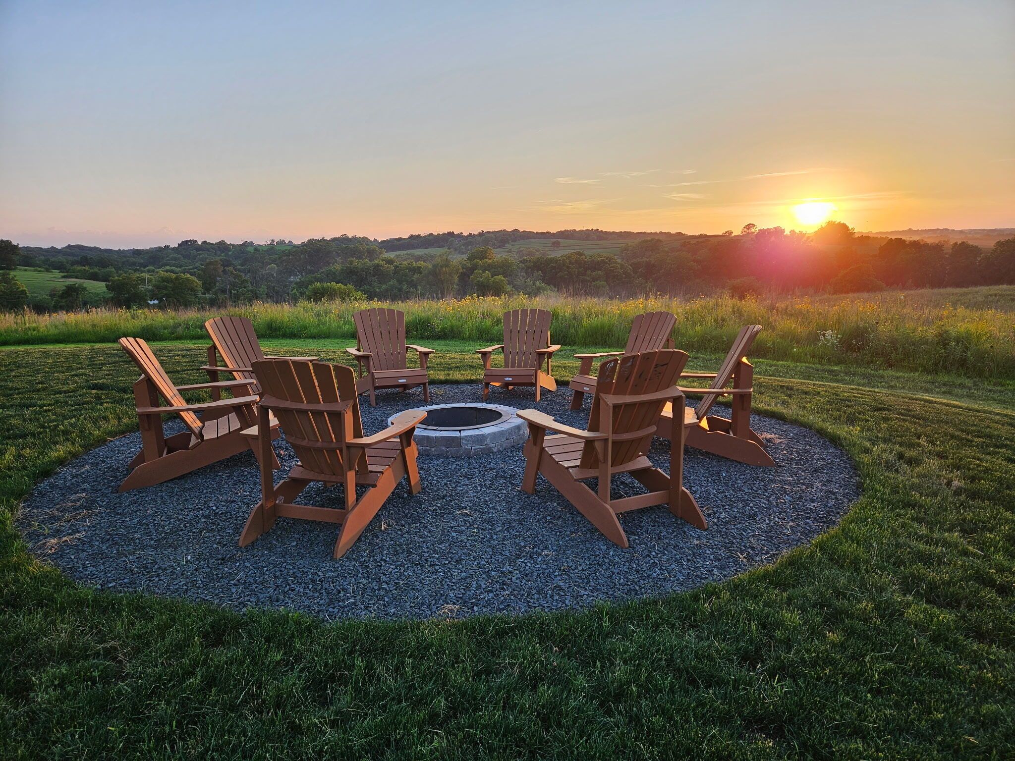 Un ensemble de chaises Adirondack en bois disposées en cercle autour d’un foyer central, lui-même situé au milieu d’un espace recouvert de gravier, le tout surmonté d’un magnifique coucher de soleil sur un champ verdoyant.