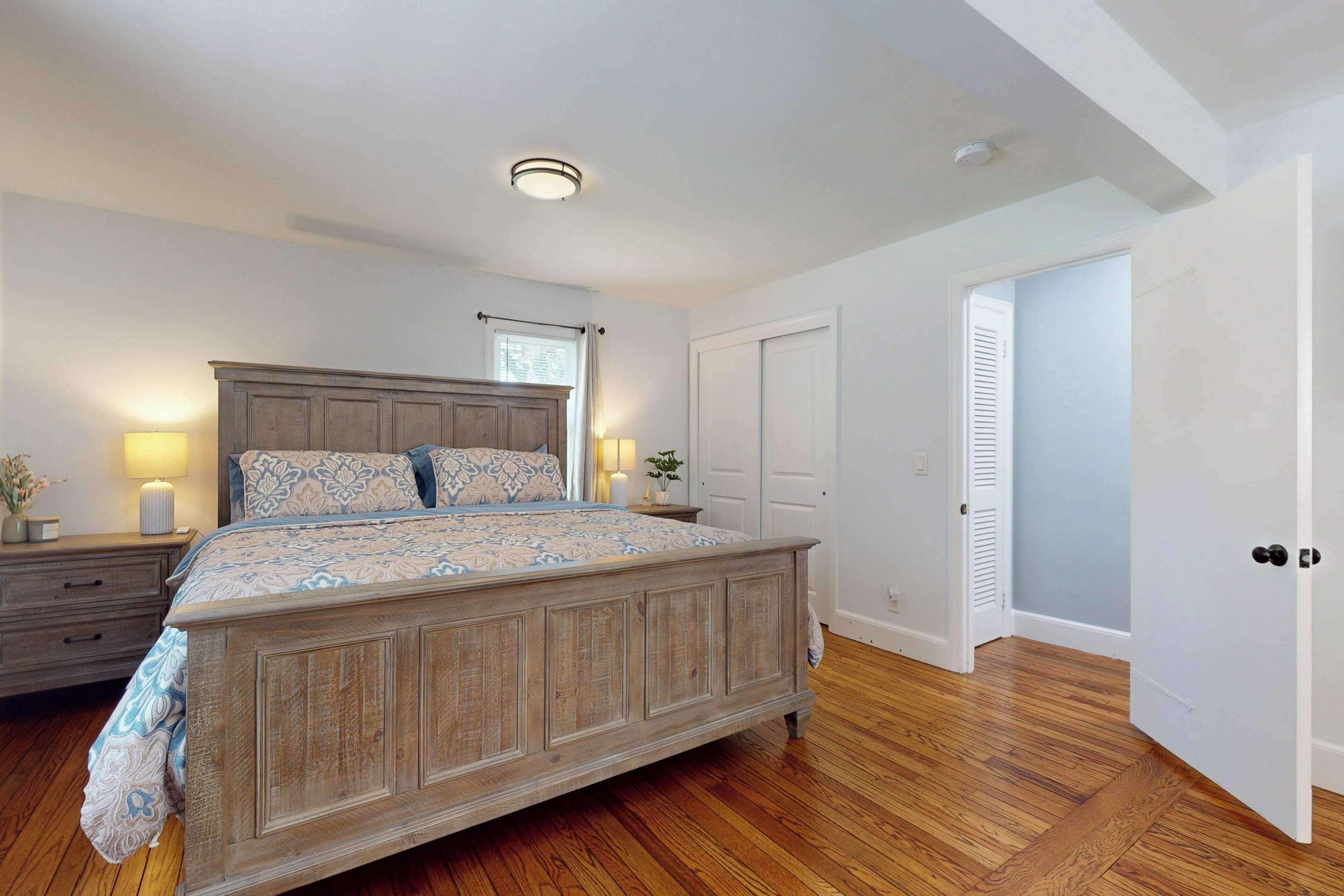 A bedroom with a wooden bed, a nightstand, a lamp, and a window with curtains.