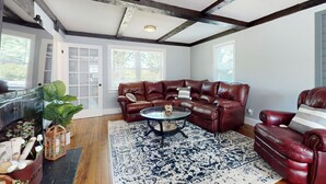 Un salon avec un canapé en cuir, une table basse en verre et un tapis à motifs. Le plafond est en bois, avec des poutres apparentes, et une porte donne sur l’extérieur.