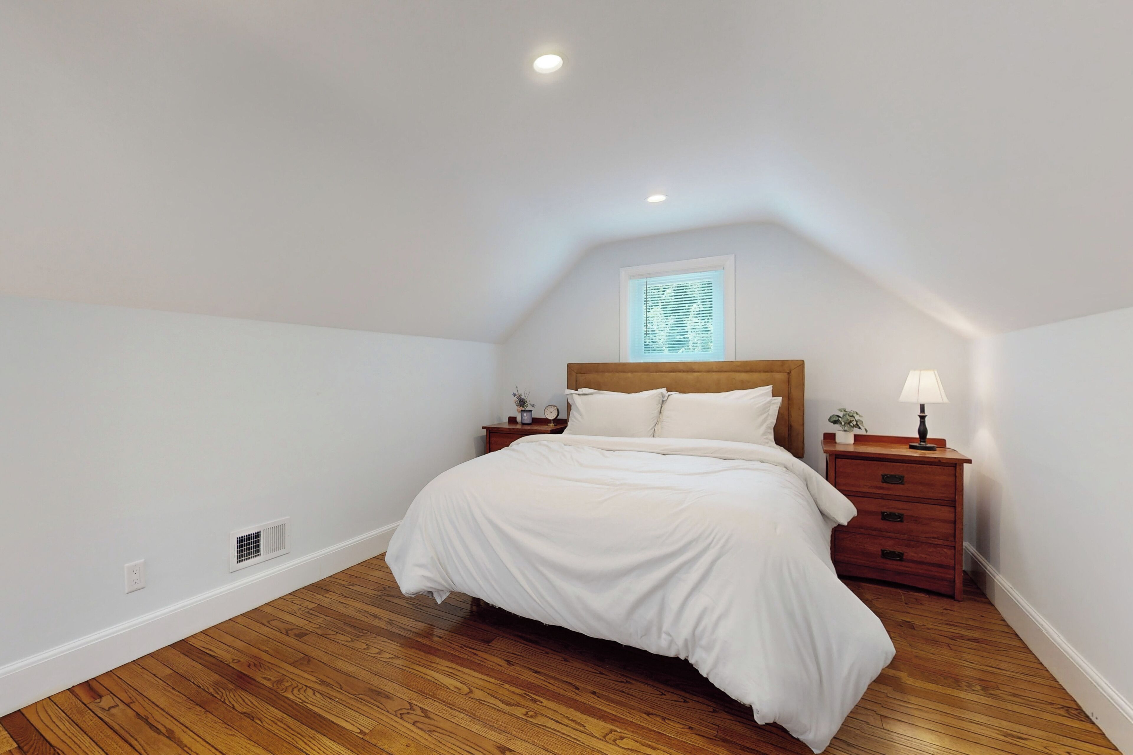 Een slaapkamer met een houten vloer, een bed met wit beddengoed, een nachtkastje met een lamp en een raam met jaloezieën.