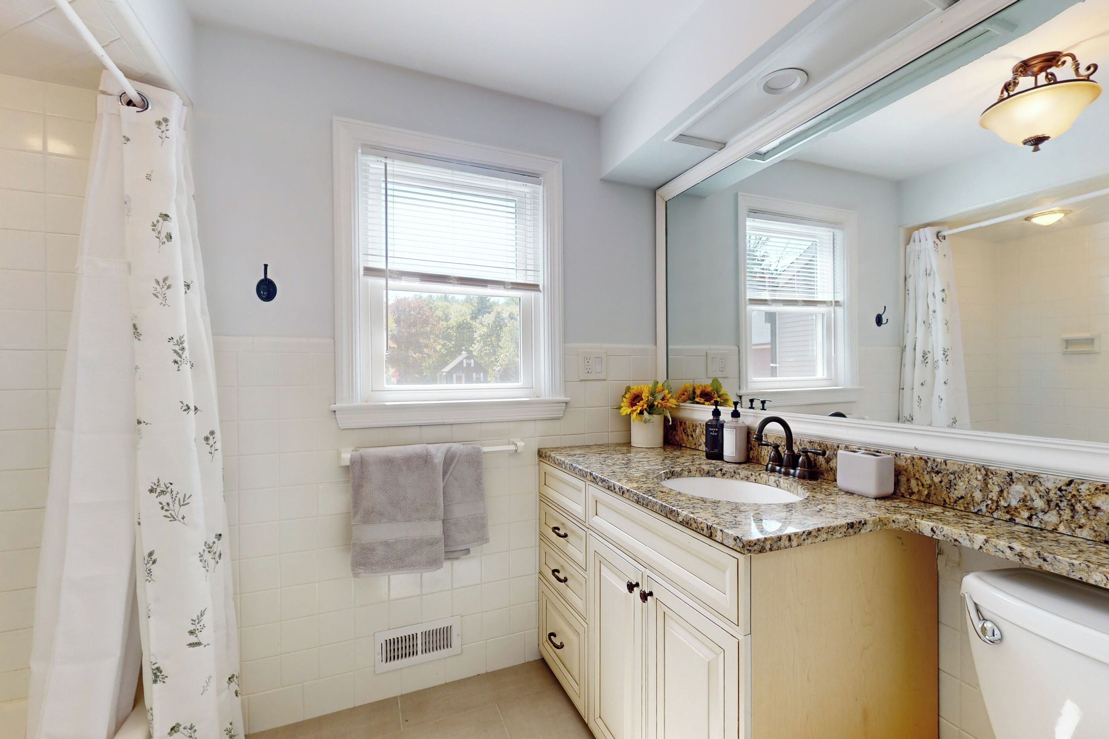 A bathroom with a white tiled floor, a sink with a granite countertop, a toilet, and a shower with a floral shower curtain. There are two windows with blinds, a mirror above the sink, and a towel rack with a towel hanging on it.