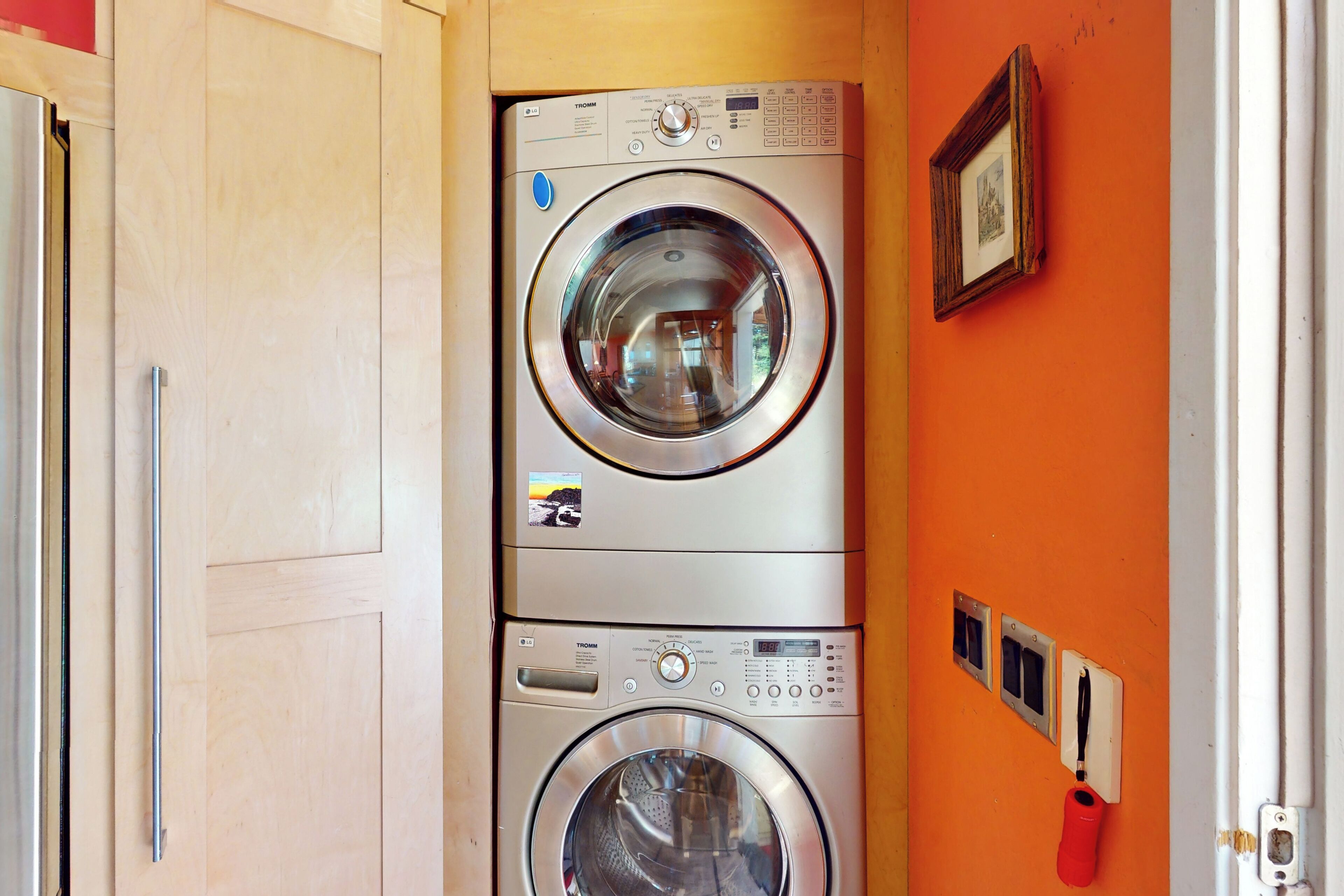 Un appareil combiné lave-linge et sèche-linge, de type « superposé », avec une porte en bois et un mur orange.