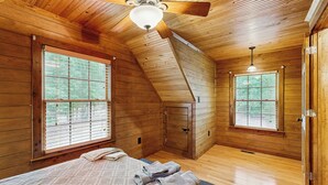 Una habitación de cabaña de madera con una cama, dos ventanas, un ventilador de techo y un armario de madera.