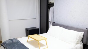 Ein ordentlich bezogenes Bett mit einer grauen Decke, einem weißen Kopfkissen und einem kleinen hölzernen Hocker.